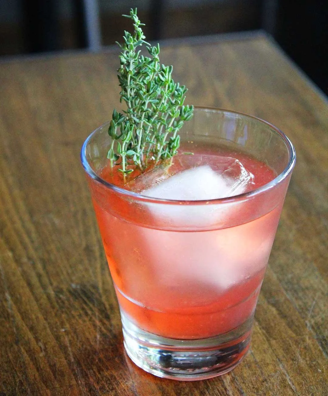 Pink cocktail with herb sprig and large ice cube in a rocks glass on wood