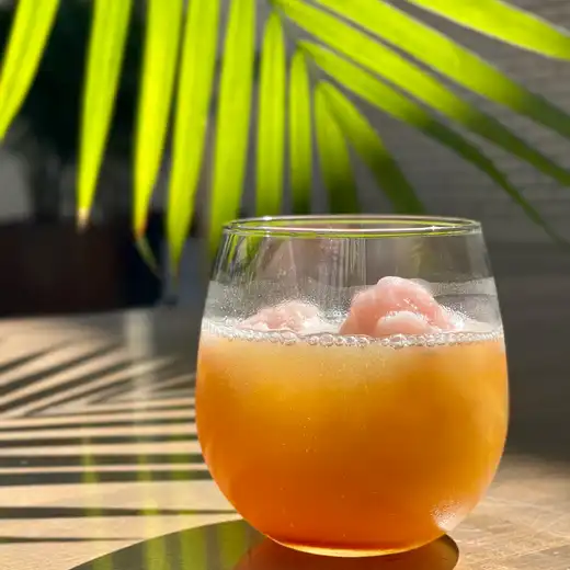 Amber cocktail in a stemless glass on a wooden table with palm fronds behind