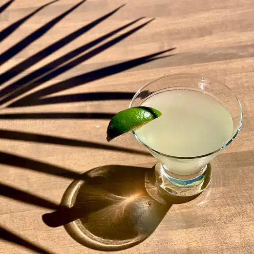 Pale yellow cocktail with a lime wedge in a small stemmed glass on a wooden table
