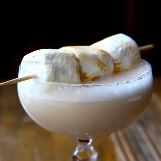 Coupe glass with foamy cream-colored cocktail topped with toasted marshmallows on a skewer