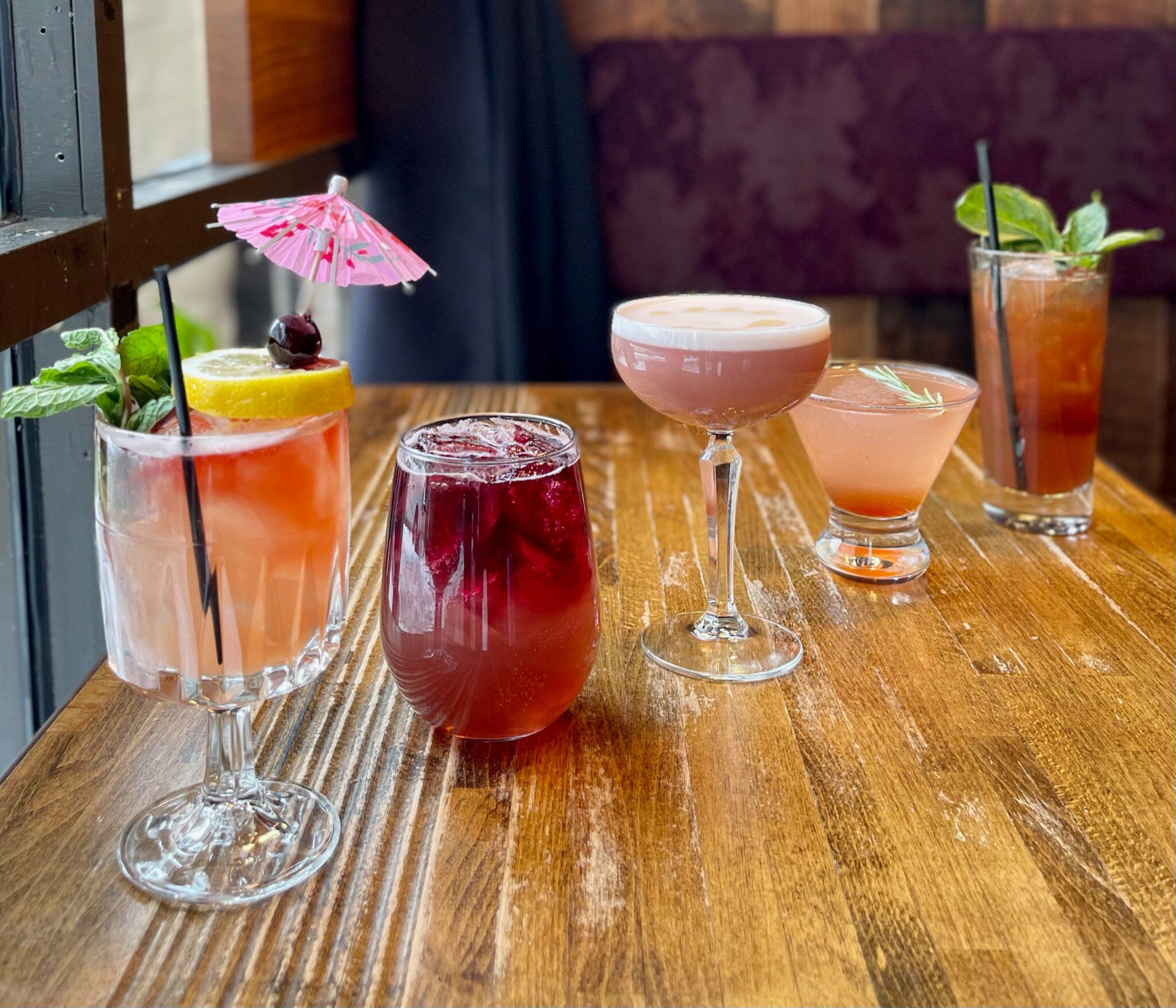 Five varied cocktails in different glasses lined up on a wooden table