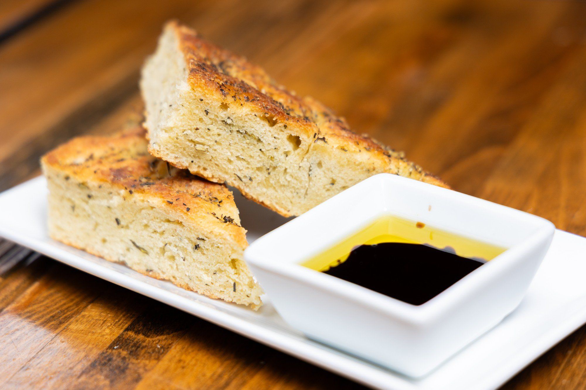 Two pieces of herbed focaccia on a white plate with a small bowl of oil and balsamic