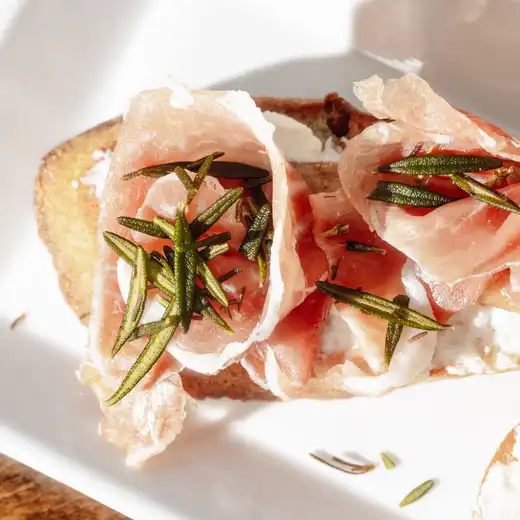Crostini topped with cured meat, white cheese and rosemary on a white plate
