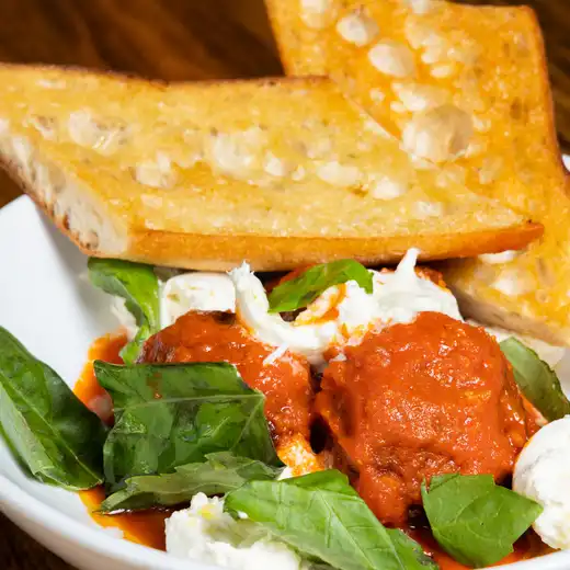 Bowl of meatballs in red sauce with white cheese, basil leaves and toasted bread slices