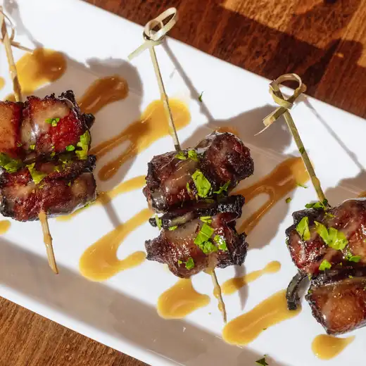 Three glazed meat skewers garnished with parsley and amber sauce on a white rectangular plate