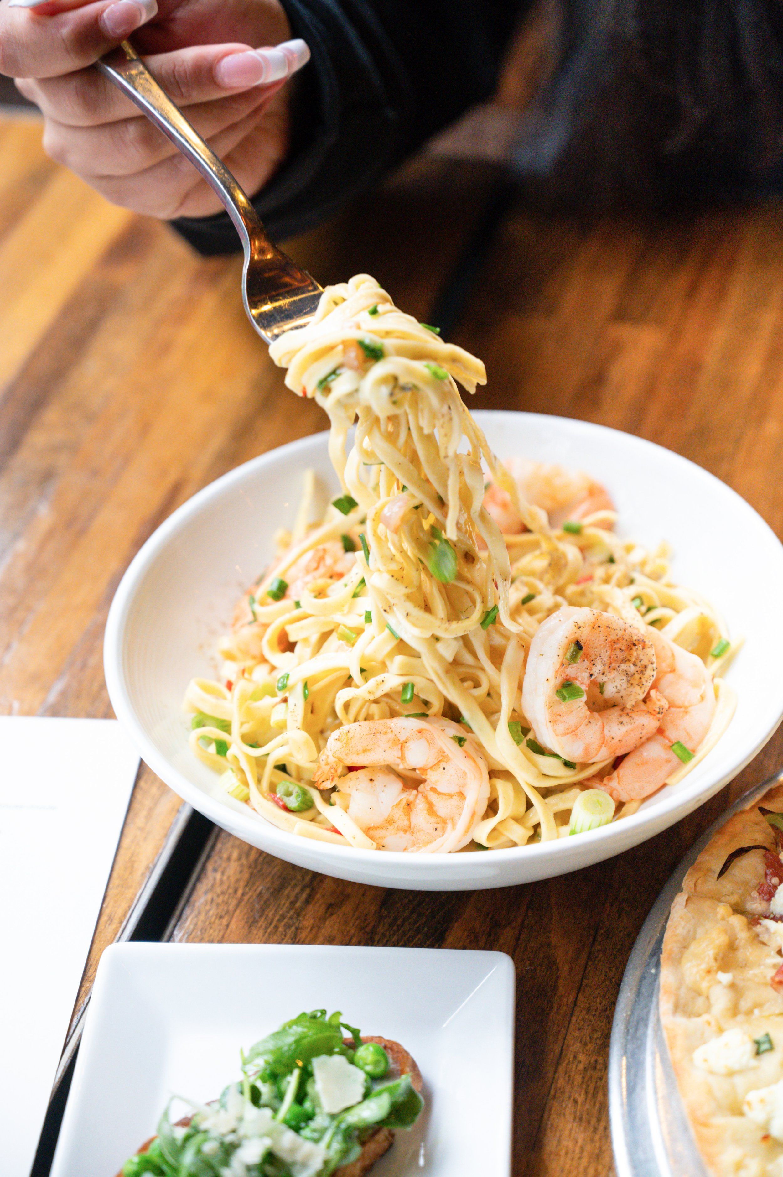 Hand lifting a fork of pasta with shrimp from a bowl, with a crostini and pizza visible on the table