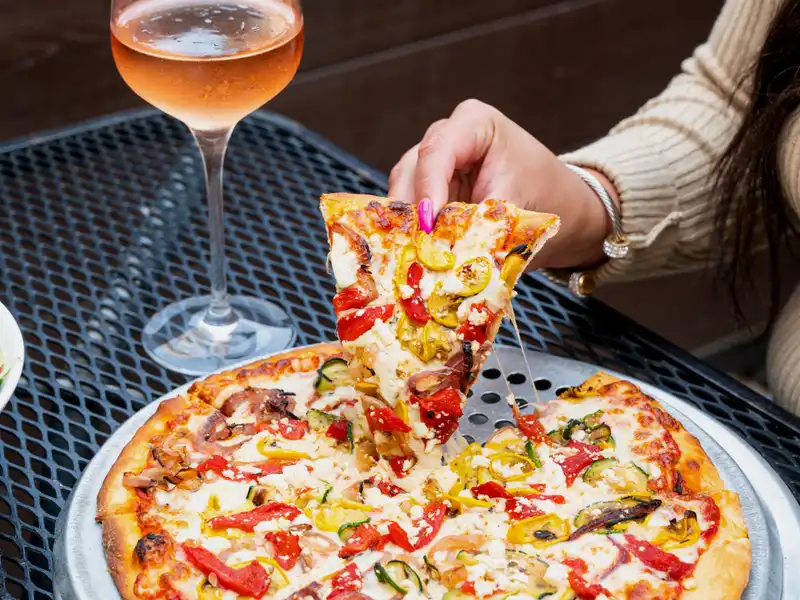 Hand lifting a slice of vegetable pizza beside a glass of pink wine on a patio table