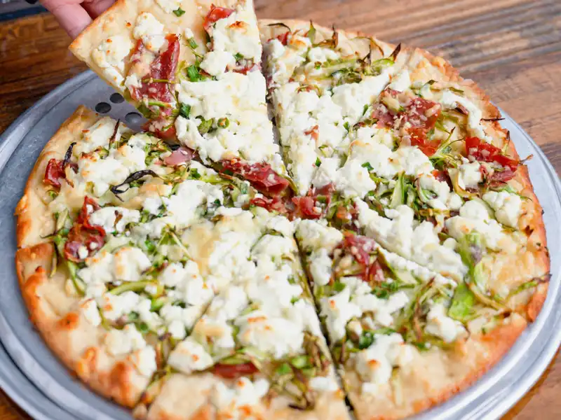 Hand lifting a slice of pizza topped with crumbled white cheese, greens and pickled peppers