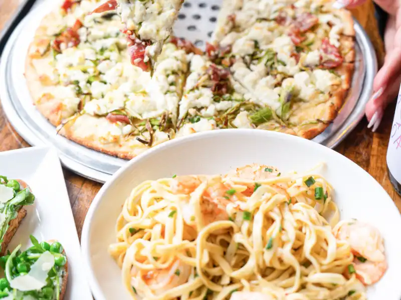 Hand lifting a slice of white pizza above a bowl of pasta with shrimp and a side crostini