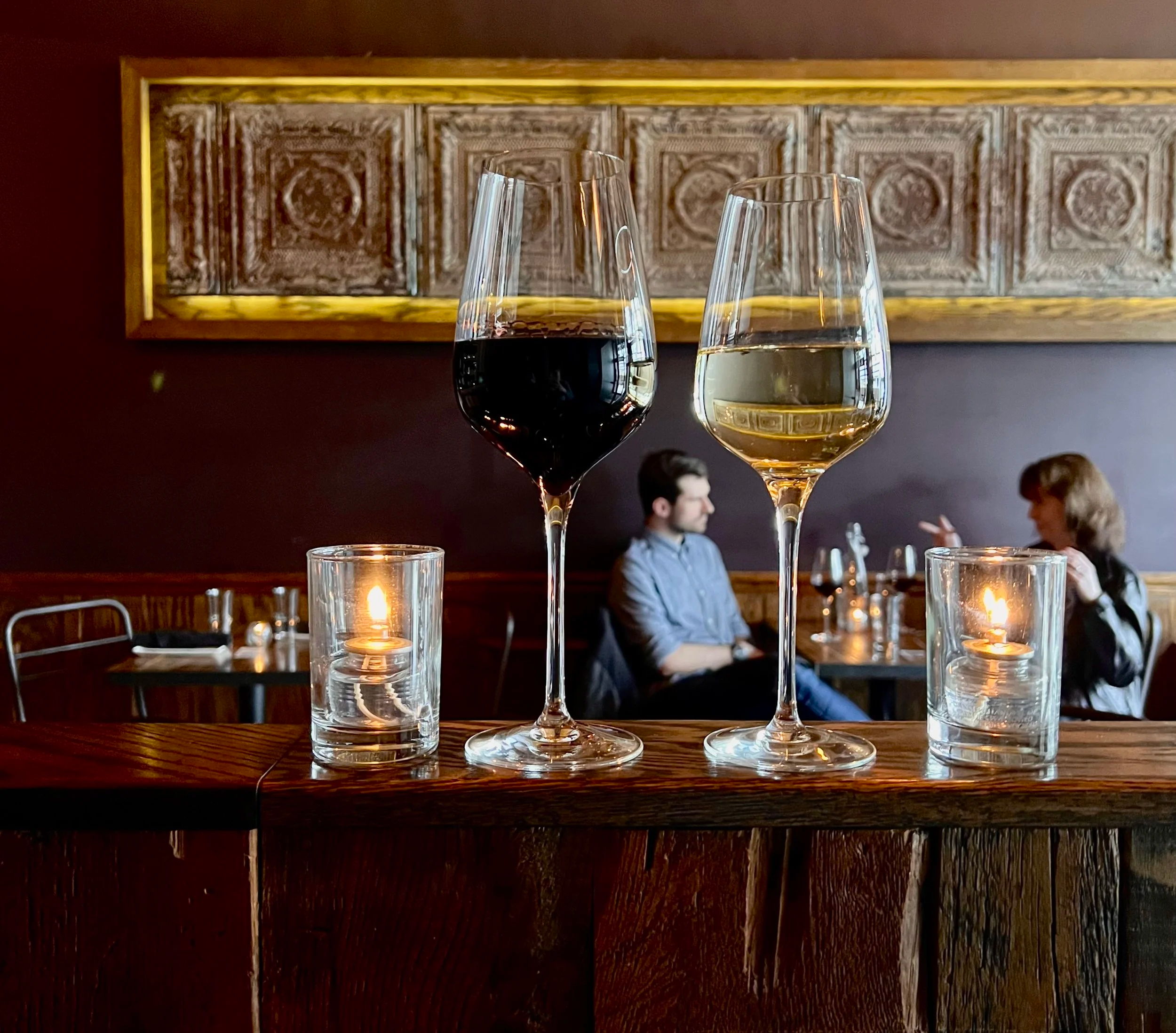 Glasses of red and white wine on a wooden bar with candles, diners behind