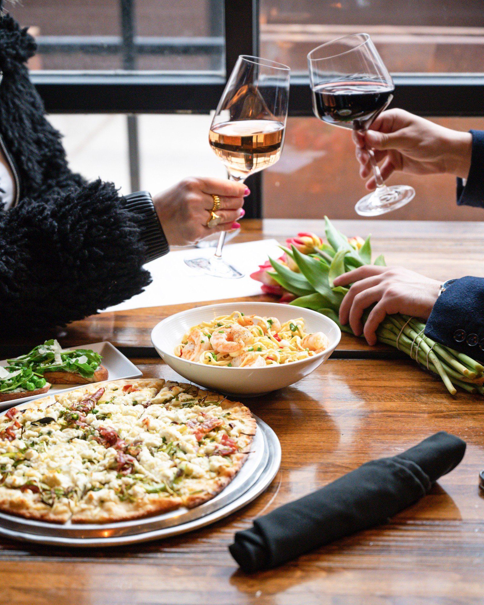 Pizza, bowl of pasta with shrimp, wine glasses, and salad on a wooden table