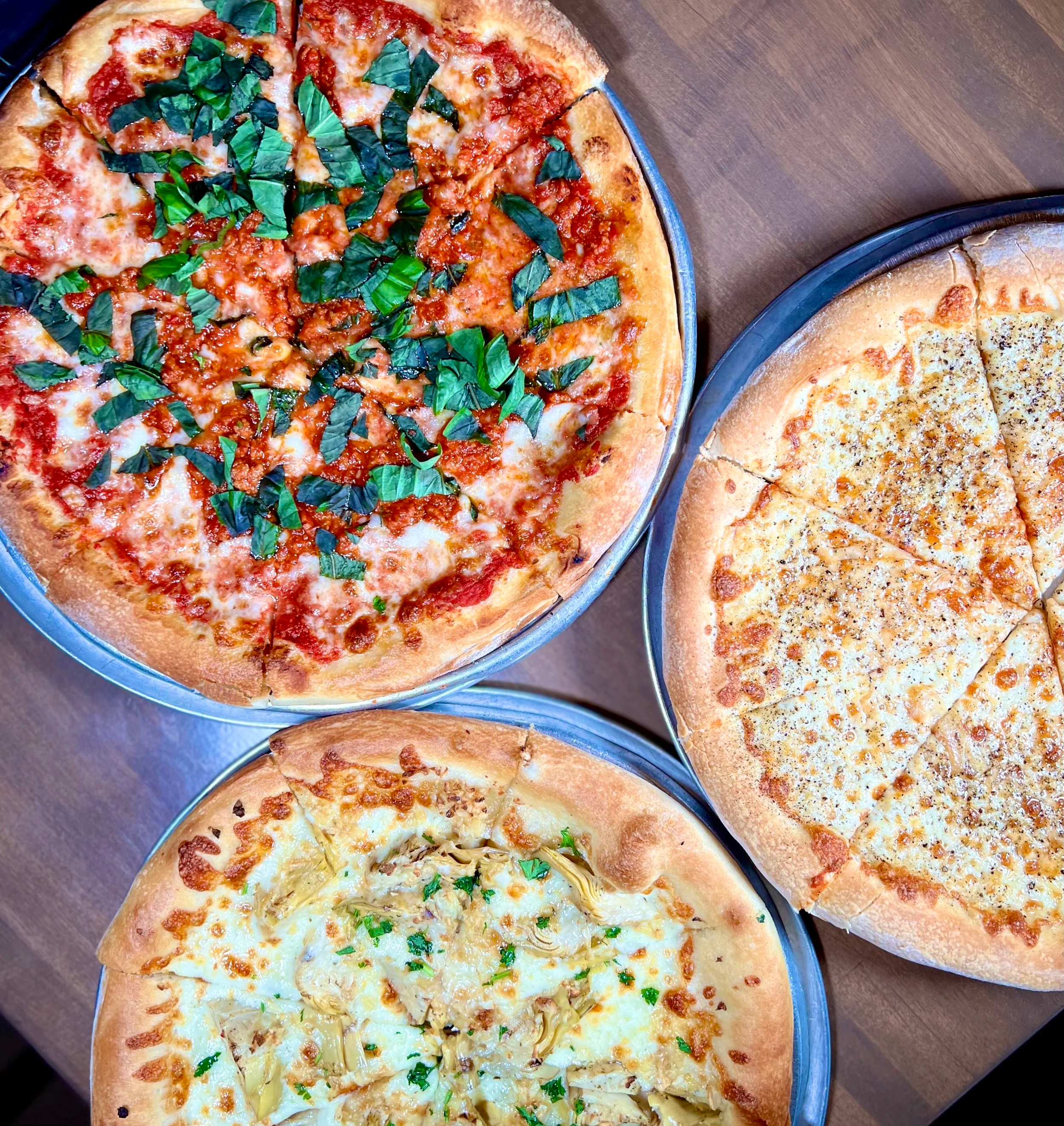 Three pizzas on metal pans on a wooden table, viewed from above