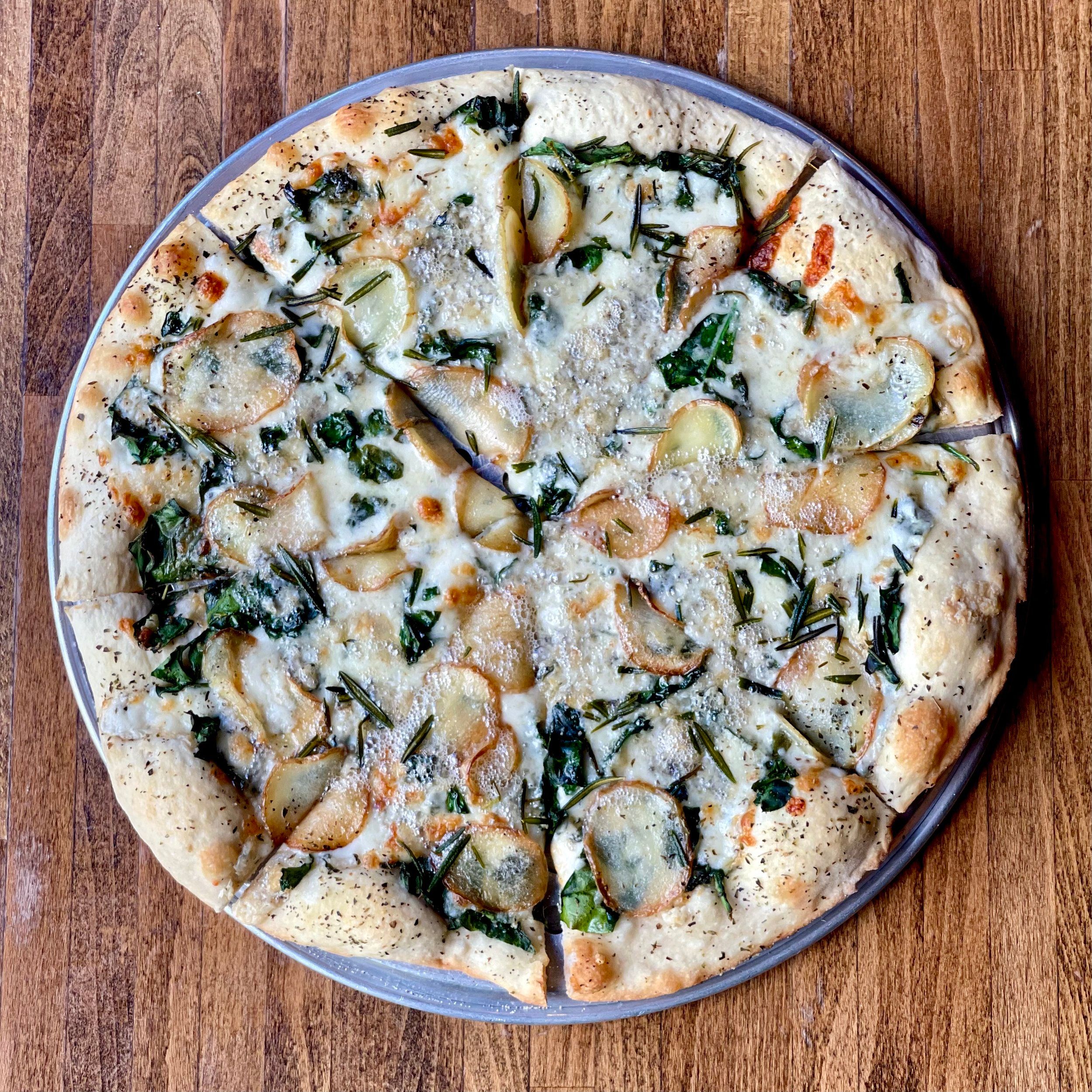 Round pizza on a metal pan topped with white cheese, sliced potatoes, and rosemary sprigs