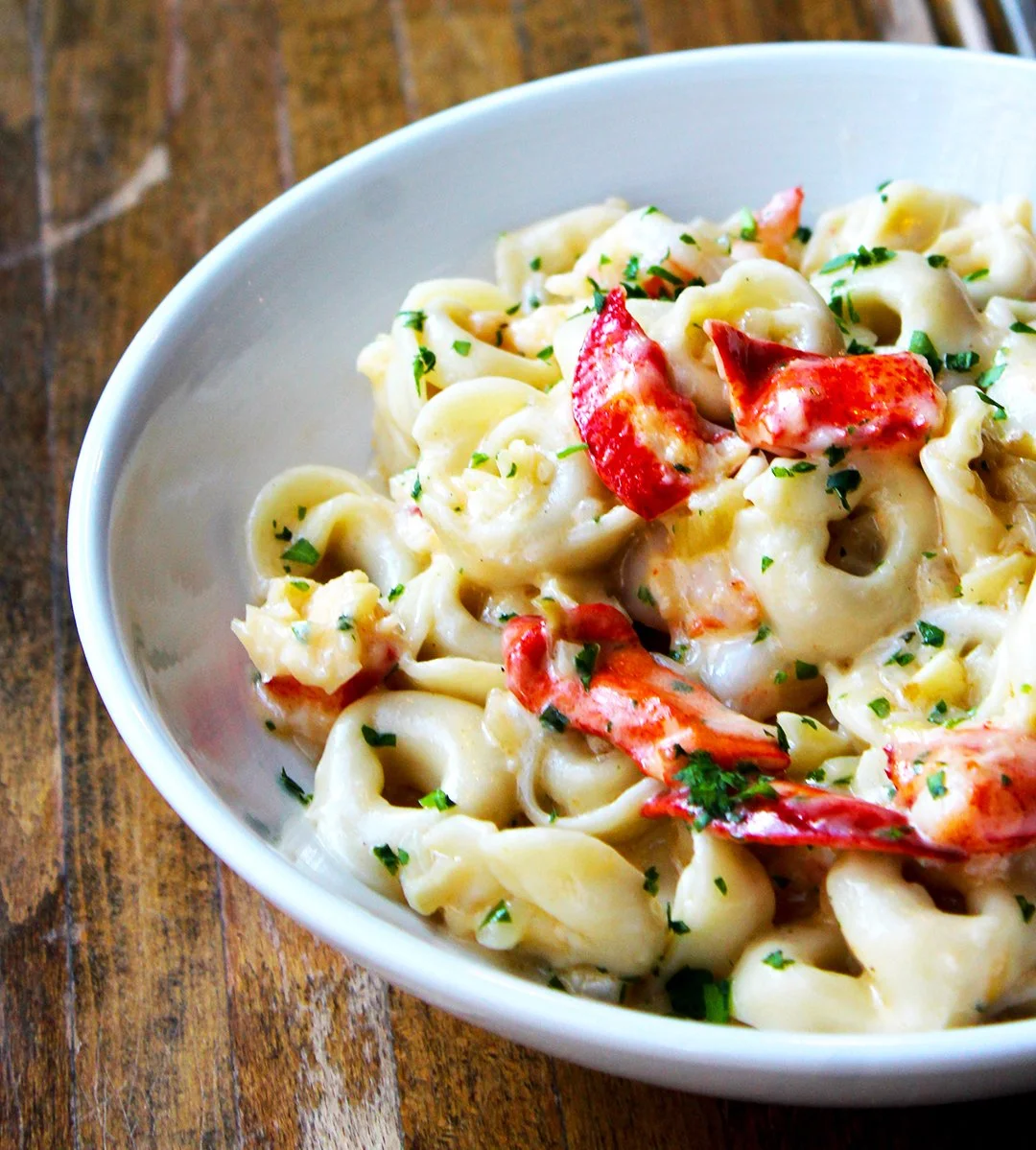 Tortellini in cream-colored sauce with red lobster pieces and parsley in a white bowl