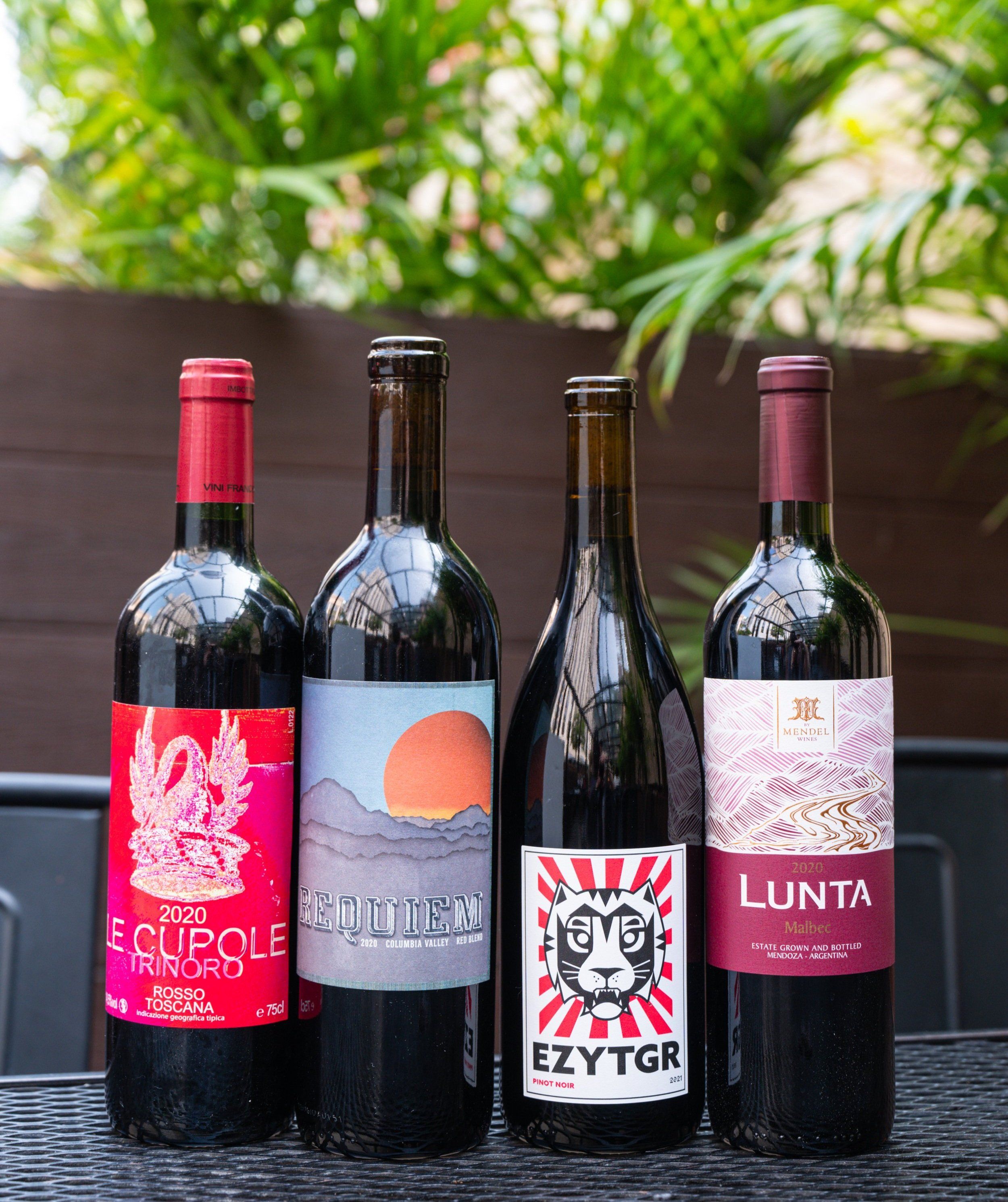 Four wine bottles with colorful labels lined up on a patio table with greenery behind