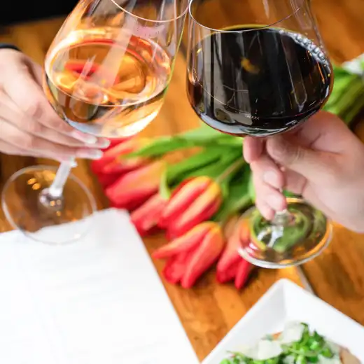 Two hands clinking glasses of pink and red wine over a wooden table with tulips and crostini