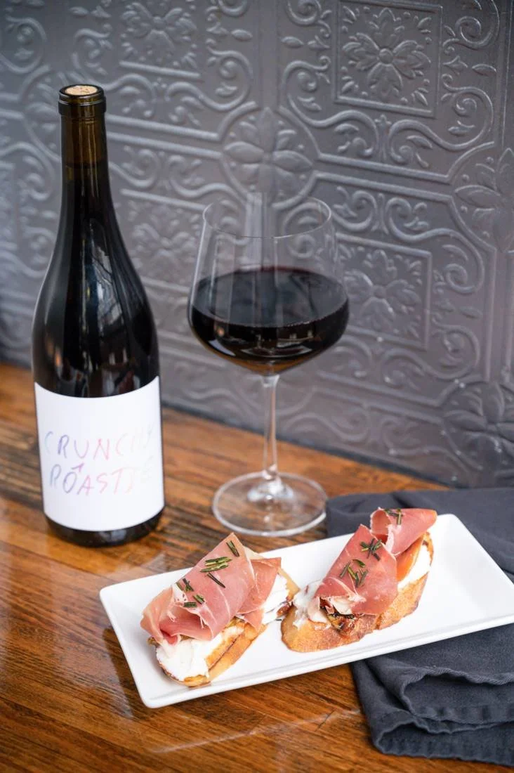 Two crostini with white cheese and cured meat beside a wine bottle and glass of red wine