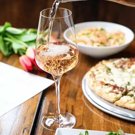 Pink wine being poured into a glass with crostini, pasta bowl, and pizza on a wooden table
