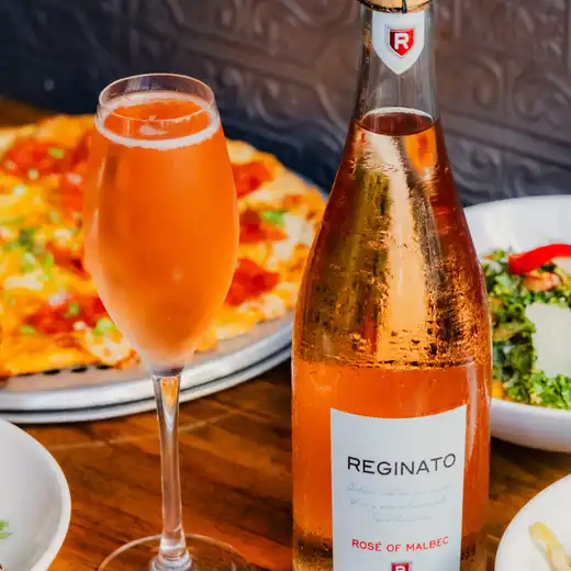 Bottle of pink sparkling wine with a flute of pink drink, pizza, and salad on a wooden table